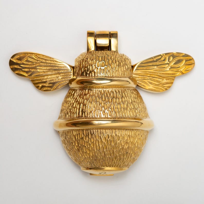 K&ouml;nnte beinhalten: Ein goldfarbener T&uuml;rklopfer aus Metall in Form einer Hummel. Die Hummel hat einen strukturierten K&ouml;rper mit einem glatten, polierten horizontalen Band in der Mitte und detaillierten, fl&uuml;gelartigen Strukturen, die vom Scharniermechanismus ausgehen.