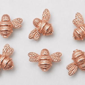 Puede incluir: Seis dijes en forma de abeja de color oro rosa dispuestos en dos filas. Cada dije presenta un cuerpo y alas texturizados con bandas horizontales, un acabado metálico y una anatomía detallada con una cabeza y abdomen redondeados.