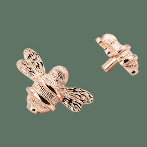 May include: Two rose gold bee-shaped drawer pulls. The bee is detailed with textured wings and a body.