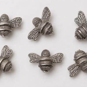 May include: Six individual silver-toned metal bee-shaped buttons or charms. Each piece features a detailed, textured body with horizontal stripes and embossed wings. The bees are arranged in two rows of three, showcasing a metallic finish.