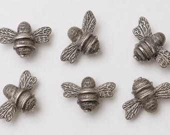 May include: Six individual silver-toned metal bee-shaped buttons or charms. Each piece features a detailed, textured body with horizontal stripes and embossed wings. The bees are arranged in two rows of three, showcasing a metallic finish.