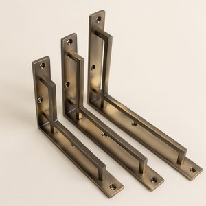 Peut inclure: Trois équerres de tablette en laiton antique de différentes tailles avec un design rectangulaire et une barre horizontale. Les équerres sont disposées en quinconce.