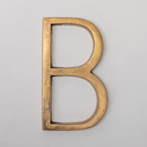 May include: A capital letter B wall sign featuring a metallic gold finish with a textured, mottled surface. The letter has a block-style font with a flat front face and straight, vertical edges, creating a three-dimensional appearance.