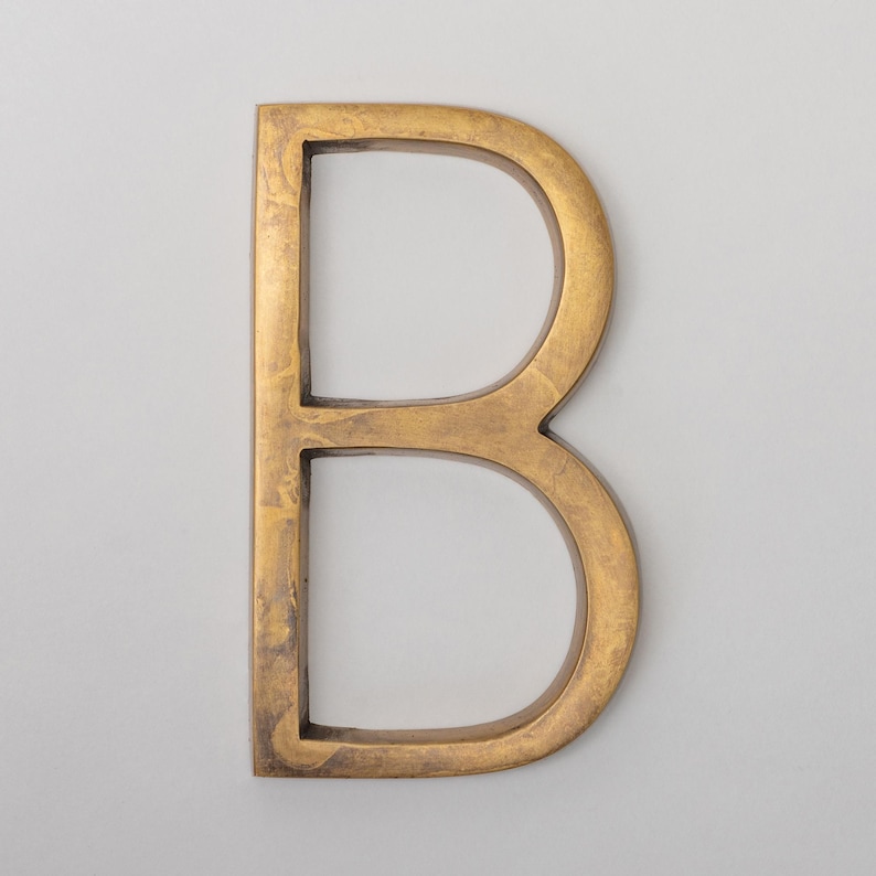 May include: A capital letter B wall sign featuring a metallic gold finish with a textured, mottled surface. The letter has a block-style font with a flat front face and straight, vertical edges, creating a three-dimensional appearance.