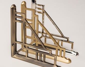 May include: Three L-shaped metal shelf brackets arranged in a row. The front bracket has an antique brass finish, the middle one is brushed gold, and the back one is polished chrome. Each bracket features a geometric openwork frame design.