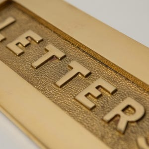 May include: Close-up of a gold-colored letter plate with the word "LETTER" embossed in capital letters. The letters are raised and have a polished finish, contrasting with the textured background.