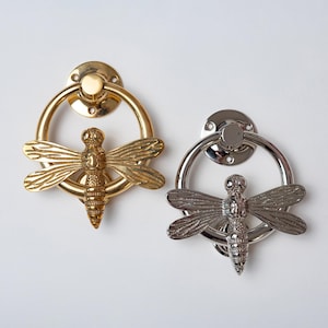 Può includere: Due battenti per porta a forma di libellula, uno con finitura color oro lucido e uno con finitura color argento lucido. Ognuno presenta una piastra di montaggio circolare, un anello e una figura di libellula dettagliata al centro.