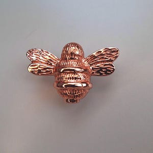 May include: A metallic copper-colored bee-shaped cabinet knob or decorative pull. The object features a shiny, reflective finish with detailed, textured wings and a segmented body, capturing the form of a bee in a polished, solid metal construction.