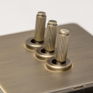 May include: A brushed brass light switch plate with three round toggle switches. Each switch has a knurled brass knob.