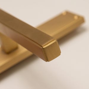 May include: A brushed brass cabinet handle featuring a rectangular bar design with beveled edges and a smooth metallic finish. The hardware includes a flat mounting plate with visible screw holes, showcasing a clean and uniform golden tone throughout.