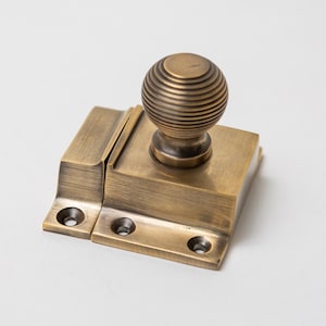 May include: A brass-toned window latch with a decorative, ridged ball knob. The latch is rectangular with screw holes for mounting. The metal has an aged appearance, suggesting a vintage or antique style.