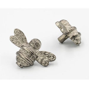 May include: Two silver bee-shaped cabinet knobs with a textured finish. The bee's body is a rounded shape with a striped pattern. The wings are detailed and spread out.