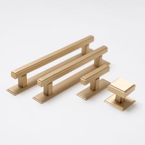 Puede incluir: Un juego de cuatro tiradores de armario de color dorado en varios tamaños. Los tiradores tienen una forma rectangular con una barra central elevada. El tirador más pequeño es un pomo.