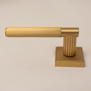 Könnte beinhalten: Ein mattgoldener Türgriff mit einem horizontalen Hebel, der eine gerippte, zylindrische Textur aufweist. Der Hebel ist mit einem vertikalen, gerippten zylindrischen Pfosten verbunden, der auf einer quadratischen, flachen Grundplatte montiert ist.