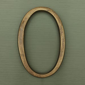 May include: A house number zero in an oval shape, featuring a metallic bronze finish with a distressed, textured surface. The numeral has a smooth, rounded profile and a consistent thickness throughout its entire circular frame structure.