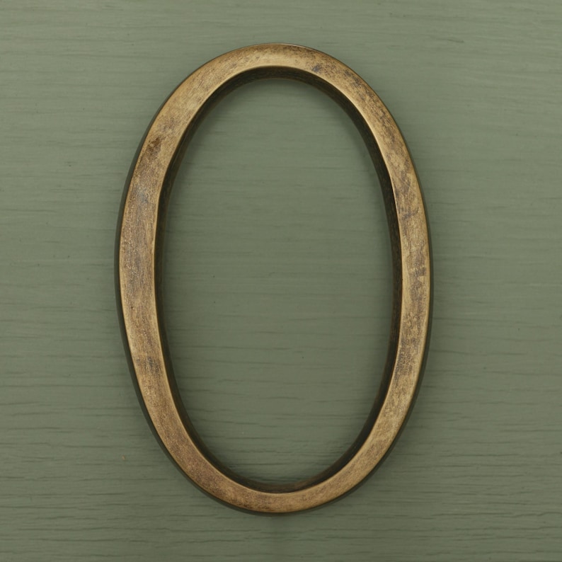 May include: A house number zero in an oval shape, featuring a metallic bronze finish with a distressed, textured surface. The numeral has a smooth, rounded profile and a consistent thickness throughout its entire circular frame structure.