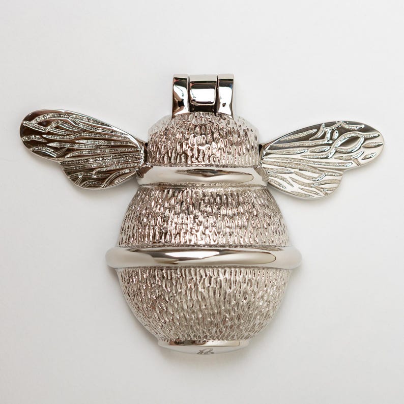 K&ouml;nnte beinhalten: Ein silberfarbener T&uuml;rklopfer in Form einer Hummel. Das Insekt hat einen strukturierten, runden K&ouml;rper mit detaillierten Fl&uuml;geln, die zur Seite ragen, und einen glatten, polierten Scharniermechanismus an der Oberseite f&uuml;r das bewegliche Teil.