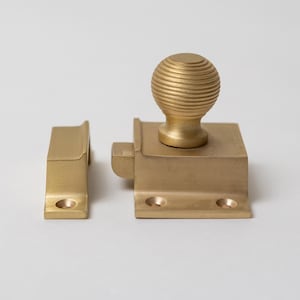 May include: A brass window sash lock set. The set includes a rectangular base with a round, ribbed knob and a matching strike plate. The hardware is a warm gold color and appears to be made of solid brass.