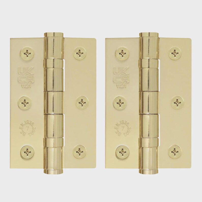 May include: Two polished brass door hinges featuring a central barrel and three screw holes on each side. The metal plates are stamped with the text UKCA 1121 and BS EN 1935 7 2002. Each hinge includes three flat-head screws installed in the plates.