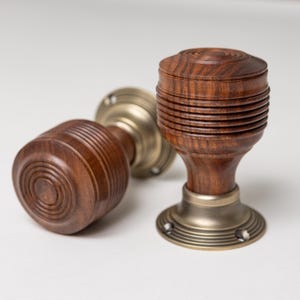 May include: Two door knobs featuring dark brown wood handles with horizontal ribbed detailing and circular tops. Each knob is attached to a circular brass-toned metal base plate with visible screw holes for mounting to a door surface.