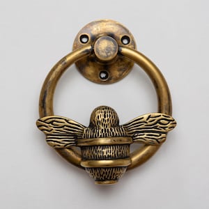 May include: A circular door knocker made of antique brass with a distressed finish. The design features a central bee figure with textured wings and body, attached to a round mounting plate with three screw holes at the top for installation.