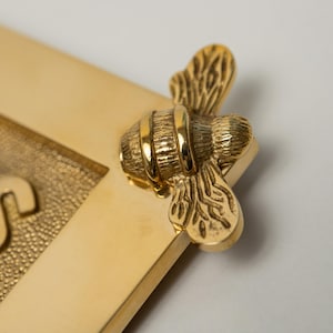 May include: A gold-colored picture frame corner with a detailed bee accent. The bee is positioned on the corner of the frame, with textured wings and body. The frame has a smooth, polished finish.