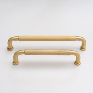 Könnte beinhalten: Zwei goldfarbene Schrankgriffe mit strukturiertem, rautenförmigem Griff. Die Griffe haben ein gebogenes Design und sind auf einer weißen Oberfläche montiert. Die Hardware ist für Schränke, Schubladen oder andere Möbel.