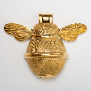 May include: A gold-colored metal door knocker shaped like a bumblebee. The bee features a textured body with a smooth, polished horizontal band across the center and detailed, wing-like structures extending from the top hinge mechanism.