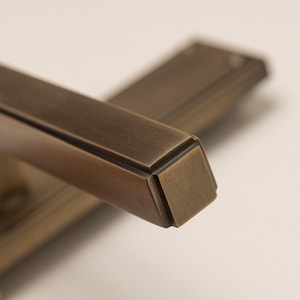 Könnte beinhalten: Ein Paar rechteckige Türgriffe mit gebürstetem Bronze-Finish. Jeder Griff zeichnet sich durch ein lineares Design mit scharfen, definierten Kanten und einer glatten, metallischen Oberfläche aus. Die Griffe sind schräg angeordnet und zeigen das quadratische Profil.