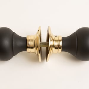 Może przedstawiać: Para czarnych gałek drzwiowych z mosiężnym wykończeniem. Gałki są okrągłe i mają gładką powierzchnię.