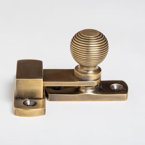May include: A brass window latch featuring a ridged ball handle. The latch is made up of rectangular and curved components, all finished in a warm, antique brass. This hardware is designed for both functionality and visual appeal, suitable for windows.