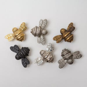 May include: Six bee-shaped metal cabinet knobs arranged in a group. The set includes one polished gold, one brushed silver, one antique bronze, one matte black, one bright chrome, and one dark gunmetal finish, each featuring detailed wing textures.