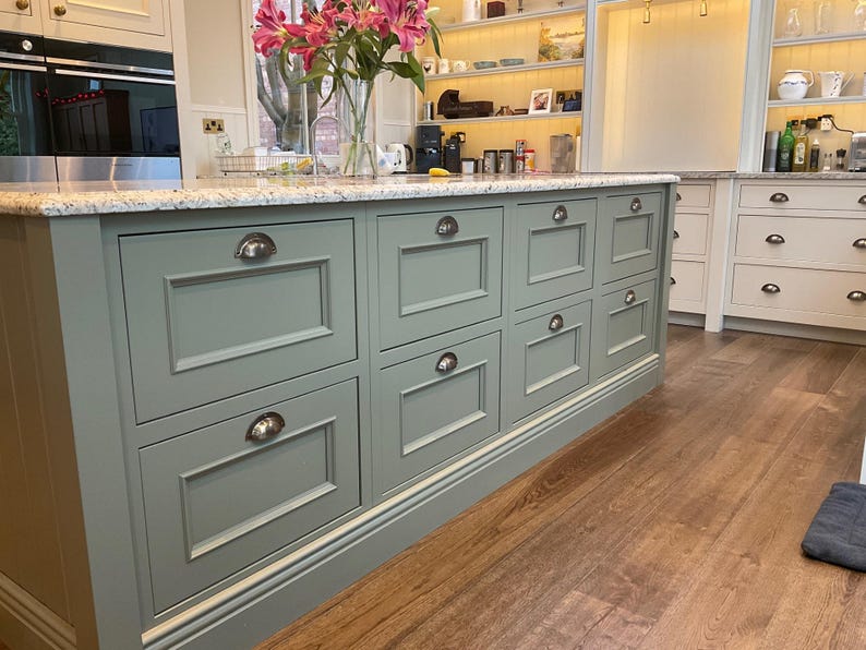 May include: A kitchen island with a light green painted finish and multiple drawers with silver handles. The island has a granite countertop and sits on a wood floor.