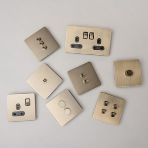 May include: A collection of eight brass light switch plates and electrical outlets. The plates are in various configurations, including single, double, and triple switches, as well as a single outlet. The plates are all made of a brushed brass finish.