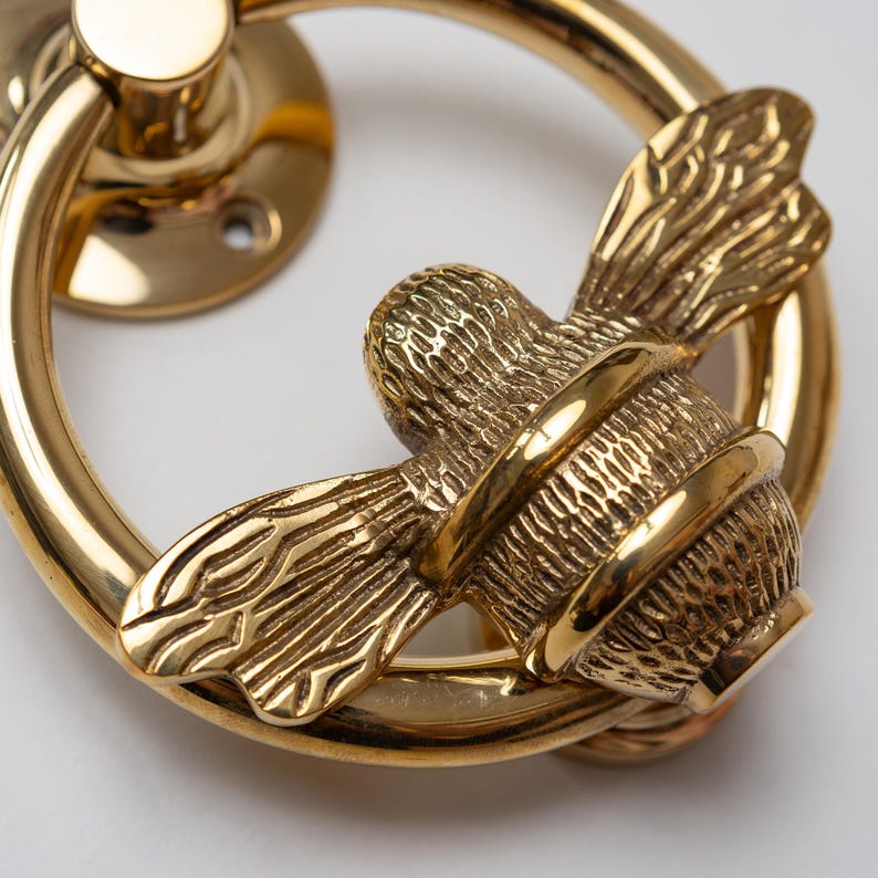 K&ouml;nnte beinhalten: Ein T&uuml;rklopfer aus poliertem, goldfarbenem Metall mit einem kreisf&ouml;rmigen Ring und einem zentralen Bienenornament. Die Biene weist detaillierte, texturierte Fl&uuml;gel und einen segmentierten K&ouml;rper auf, w&auml;hrend der Ring glatt und reflektierend ist.