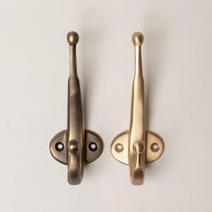 May include: Two wall-mounted coat hooks with a dual-prong design. One hook features a dark, brushed bronze finish, while the other has a lighter, satin brass finish. Each hook has an oval base plate with two screw holes for mounting purposes.