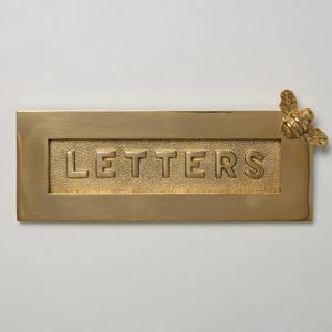 May include: A rectangular brass letterbox with the word "LETTERS" embossed on the front. A decorative bee is attached to the right side. The letterbox is mounted on a white surface.