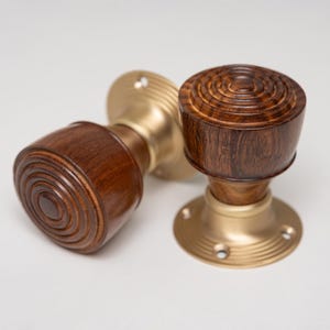 Może przedstawiać: Dwa drewniane gałki do drzwi z ciemnobrązowym wykończeniem i detalami w postaci koncentrycznych okręgów. Gałki są zamontowane na podstawach w kolorze mosiądzu z otworami na śruby, na białym tle. Gałki są prawdopodobnie do drzwi wewnętrznych.