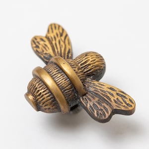 Puede incluir: Un pomo de mueble de metal en tono latón con forma de abeja. La figura del insecto presenta un cuerpo texturizado con alas detalladas y una banda lisa y elevada que rodea la sección media. El acabado tiene un aspecto oscuro y antiguo.