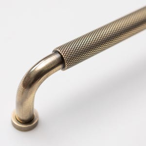 May include: Close-up of an antique brass cabinet pull. The handle features a textured, diamond-patterned grip and a curved design. The hardware has a vintage aesthetic, suitable for kitchen or furniture upgrades.