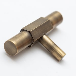 May include: A brushed brass cabinet knob with a hexagonal shape and a T-shaped design. The knob has a smooth, slightly textured finish.