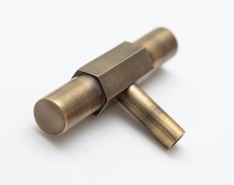 May include: A T-shaped cabinet knob with a brushed antique brass finish. The handle features a central hexagonal section connected to two cylindrical ends, with a single cylindrical post extending downward for mounting purposes.