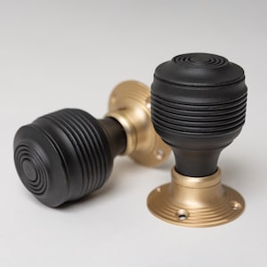 Może przedstawiać: Dwa czarne uchwyty do drzwi z żebrowanym wzorem i podstawami w kolorze mosiądzu. Gałki mają okrągły wzór na górze i są umieszczone na białym tle. Podstawy mają okrągły wzór.