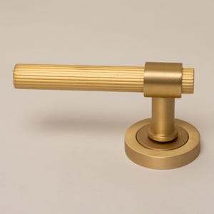 May include: A brushed gold door handle featuring a horizontal cylindrical lever with a ribbed, textured surface. The lever connects to a smooth, circular mounting plate via a short, solid cylindrical post, creating a cohesive metallic hardware piece.
