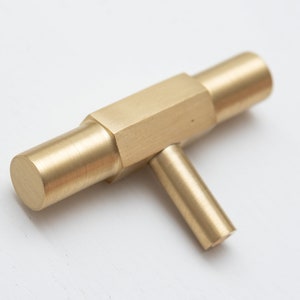 May include: A brass T-shaped cabinet knob with a square centre and rounded edges.