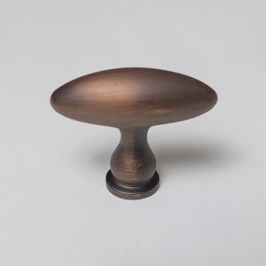 May include: A cabinet knob with an elongated, oval-shaped top and a rounded, pedestal-style base. The hardware features a dark, brushed bronze finish with visible metallic texture and subtle highlights across the curved surface.