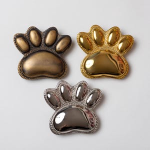 May include: Three paw print shaped metal dishes in different finishes. One is a matte antique bronze, one is a polished bright gold, and one is a polished silver. Each dish features a textured border around the smooth, raised paw pads.