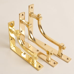 Könnte beinhalten: Vier goldene Metall-Regalböcke in verschiedenen Größen. Die Böcke sind L-förmig mit einem dekorativen, quadratischen Design an der Ecke.