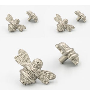 Puede incluir: Cinco pomos de armario en forma de abeja plateados con un acabado texturizado. Los pomos tienen un diseño de abeja detallado con alas, cuerpo y aguijón.