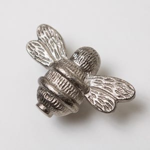 May include: A silver-toned metal cabinet knob shaped like a bumblebee. The knob features detailed, textured wings and a segmented body with horizontal ridges. The bee is depicted in a flying position with wings extended to the sides and back.